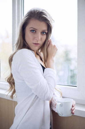 beautiful girl with a cup near the window on a light background in a white shirt and top. home photoの写真素材