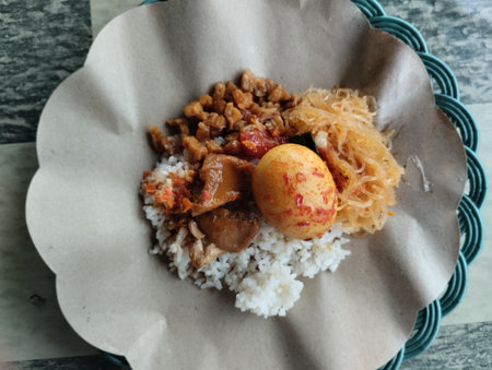 A traditional Indonesian breakfast plate featuring nasi uduk - fragrant coconut rice served with spicy boiled egg, stir-fried tempeh, and sautéed vermicelli. Presented on brown papeの写真素材