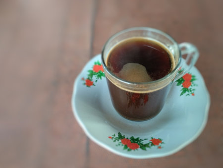 A steaming cup of black coffee served in a clear glass cup placed on a classic white saucer with red floral patterns. Captured in a warm morning atmosphere on a wooden table,の写真素材