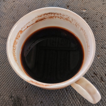 A close-up, high-angle shot of a white ceramic mug filled with rich, dark black coffee. The deep color of the coffee contrasts sharply with the white porcelain, which shows slightの写真素材
