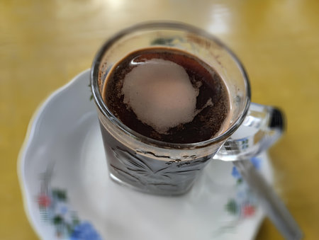 A close-up, high-angle view of a glass cup filled with hot, dark black coffee. A layer of light brown foam rests on the surface. The clear glass cup has a traditional or rusticの写真素材