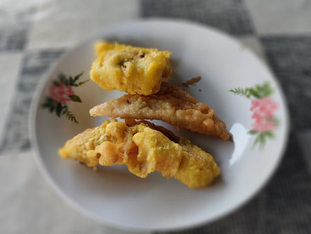 A close-up shot of freshly made banana fritters arranged on a white plate with floral decorations. The fritters are golden brown with a crunchy texture, placed on a softly blurredの写真素材