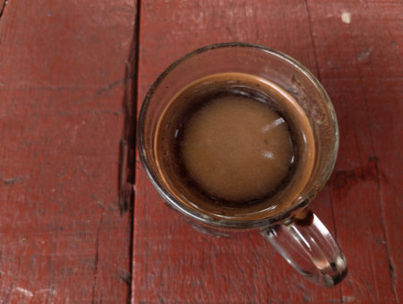 A top-down view of a hot black coffee served in a clear glass cup placed on a rustic red wooden table. The creamy layer of foam on the surface adds a warm and inviting look.の写真素材