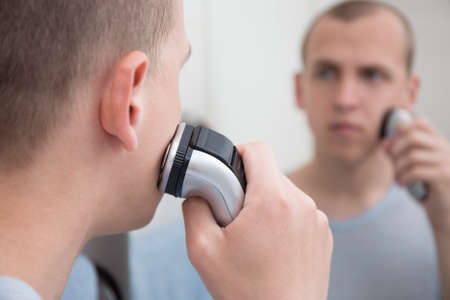 young handsome man shaving with electric razor in bathroom