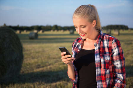 young beautiful blonde woman with mobile phone in field with haystacksの写真素材