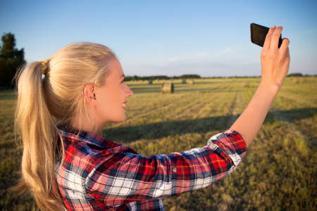 rear view of beautiful country girl making selfie photo on smartphone in field with haystacksの写真素材