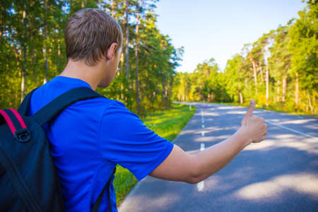 hitchhiking concept - back view of man with backpack standing on forest roadの写真素材