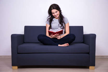 young happy woman sitting on sofa and reading bookの写真素材