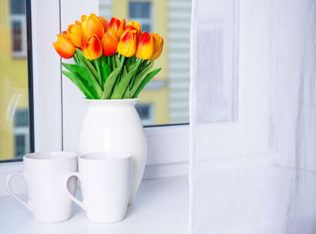 close up of beautiful orange tulips in vase and two cups on a window sill
