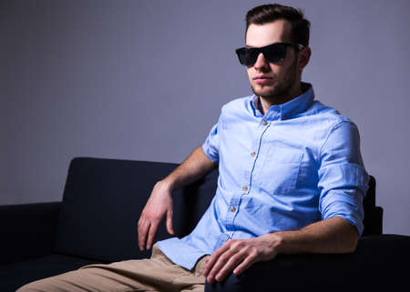 handsome man in sunglasses sitting on sofa in dark room over gray backgroundの写真素材
