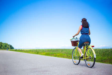 travel concept - back view of young woman riding vintage bicycle with basket in countryside