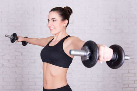 sport concept - young beautiful woman doing exercises with dumbbells over white brick wall