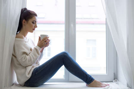 morning - young woman with cup of coffee looking out of the window at homeの写真素材