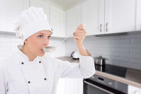 young woman in chef uniform tasting something in modern kitchenの写真素材