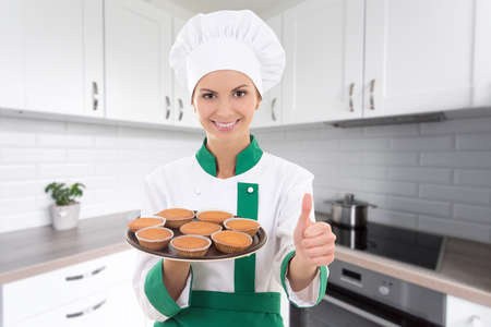 young beautiful chef woman in uniform holding tray with muffins and thumbs up in modern kitchenの写真素材