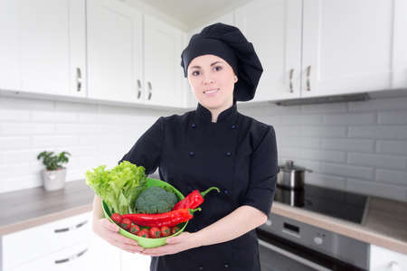 young cook woman in black uniform with bowl of vegetables in modern kitchenの写真素材