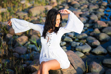 portrait of young beautiful woman in white dress sitting on rocky beachの写真素材