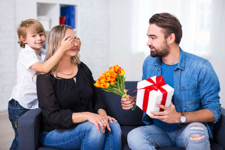 mother's or female day concept - little son and father giving flowers and gift to wife and mother at homeの写真素材