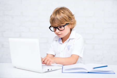 cute little school boy in glasses doing homework and using laptop in bright roomの写真素材