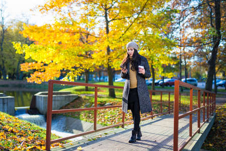 young beautiful business woman with smart phone and coffee cup walking in autumn parkの写真素材