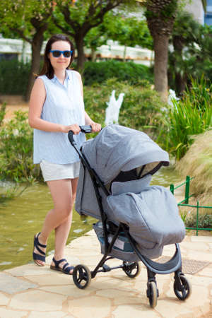 portrait of young woman walking with baby stroller in summer parkの写真素材