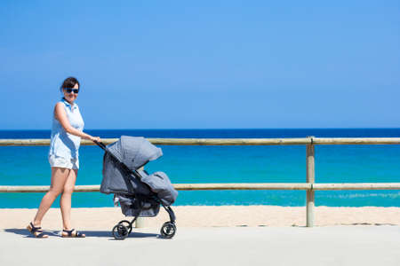 parenting and travel concept - side view of young mother pushing baby stroller on the summer beachの写真素材