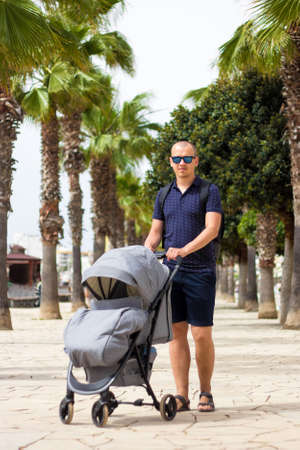 portrait of young man walking with baby stroller in summer park with palm treesの写真素材