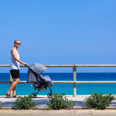 summer concept - side view of young father pushing baby stroller on the beachの写真素材