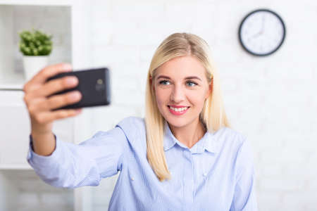 young cheerful woman taking selfie with smart phone at home or in office