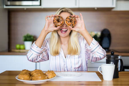 portrait of funny girl in pajamas covering her eyes with donuts in modern kitchenの写真素材