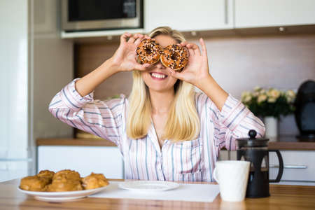 portrait of funny young woman covering her eyes with donuts in modern kitchenの写真素材