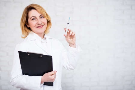 health care, vaccination and cosmetology concept - mature female doctor holding syringe and clipboard over white brick wallの写真素材