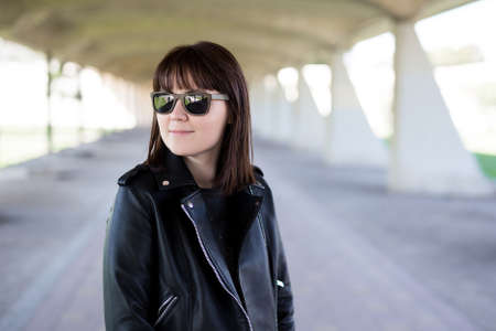 portrait of young woman in leather jacket and sunglasses posing under city bridge