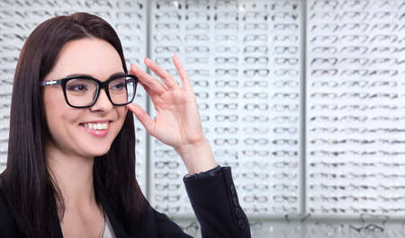 eyesight and vision concept - happy young woman trying on glasses at optics store