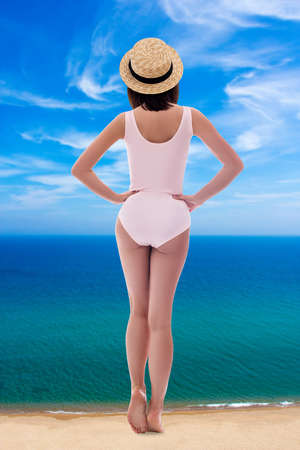 summer vacation and travel concept - back view of giant young woman in pink swimsuit and straw hat standing on sea coastの写真素材
