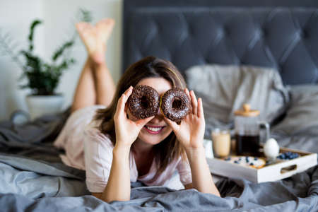 morning and breakfast concept - happy young woman in pajamas having fun in bedroom and covering her eyes with donutsの写真素材