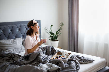 good morning and breakfast concept - portrait of happy young beautiful woman in pajamas sitting on bed and drinking coffeeの写真素材