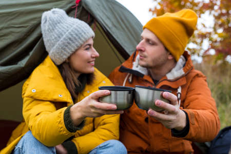 travel, trekking and hiking concept - portrait of couple drinking tea or coffee near green tent in autumn forestの写真素材