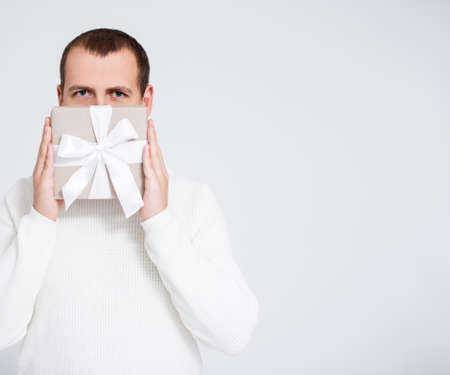 Christmas or Valentine's day concept - young man in white sweater covering face with gift box over gray background with copy spaceの写真素材