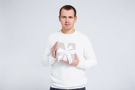 portrait of young man in warm winter sweater holding gift box over gray background with copy space