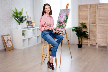 full length portrait of young woman artist with easel, palette and paint brush painting picture at...