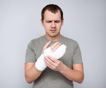 injury and first aid concept - man holding his broken hand with bandage and gypsum over gray backgroundの写真素材