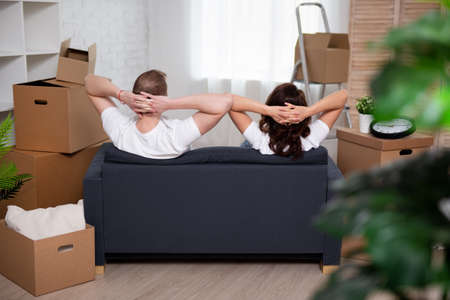 back view of happy young couple resting on couch after moving day and stack of cardboard boxesの写真素材