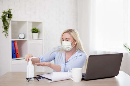 business, health care, safety and corona virus pandemic concept - young woman in mask working with laptop and using sanitizer at home or in officeの写真素材