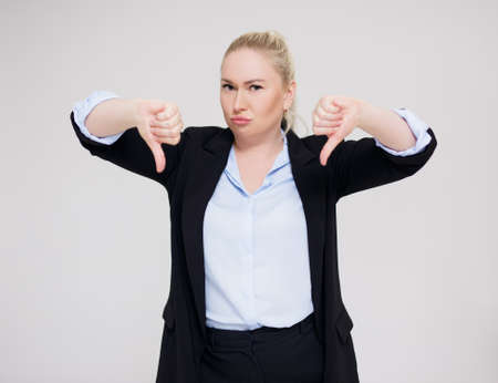 dislike - portrait of young beautiful plus size blonde woman in black business suit thumbs down over gray backgroundの写真素材