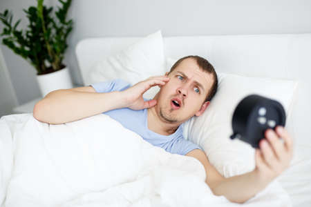 portrait of overslept shocked man looking at alarm clock lying in bed at home