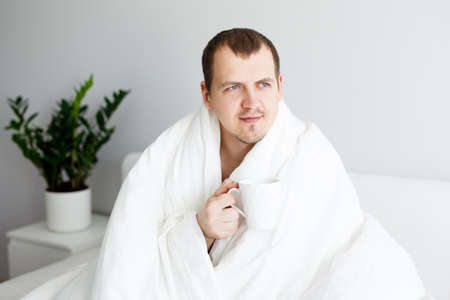 good morning concept - portrait of happy handsome man sitting on bed, wrapped in blanket and drinking coffeeの写真素材