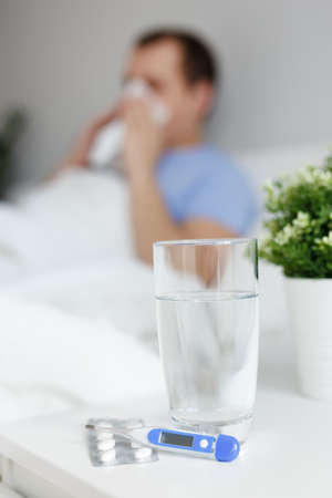 health care and illness concept - sick man lying on bed, glass of water, pills and thermometer on bedside tableの写真素材