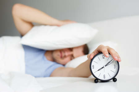 hangover and routine concept - man waking up with alarm clock and covering head with pillow in bedroom at homeの写真素材