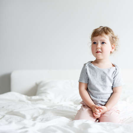cute little girl sitting on bed and dreaming or thinking about something, copy space over white wall backgroundの写真素材